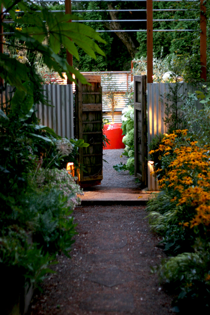 A gravel pathway is lined with lush plants leading to a doorway surrounded by lights at dusk.