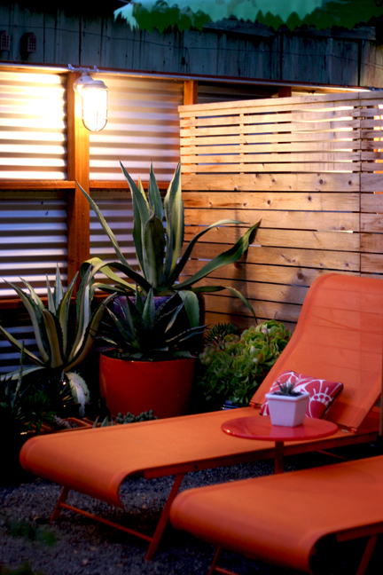 Two modern orange fabric-covered lounge chairs are nestled into a corner with exotic plants in pots against a fence.