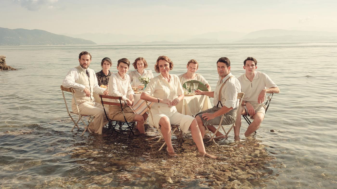 A group of people dressed in 30's white clothing sit at a table with their feet in the shallow ocean.